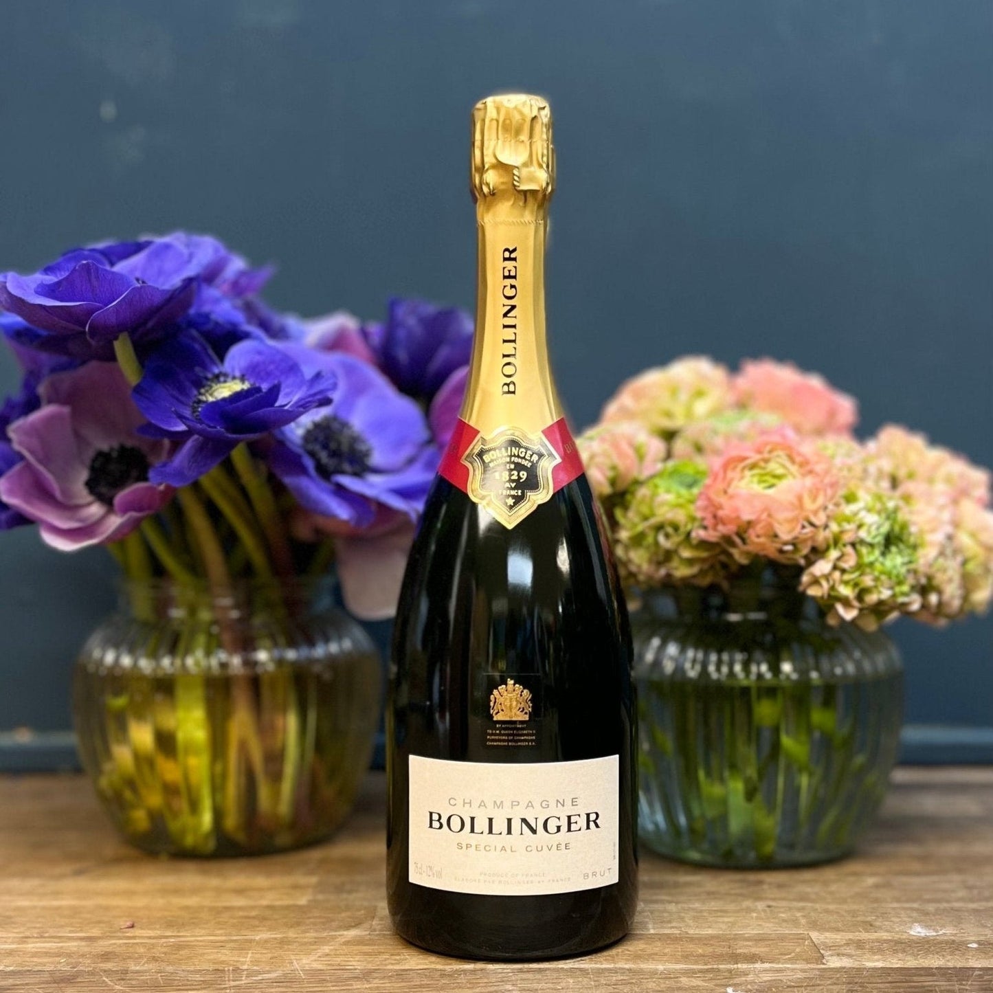 Bollinger champagne bottle with two flower arrangements on a wooden surface.