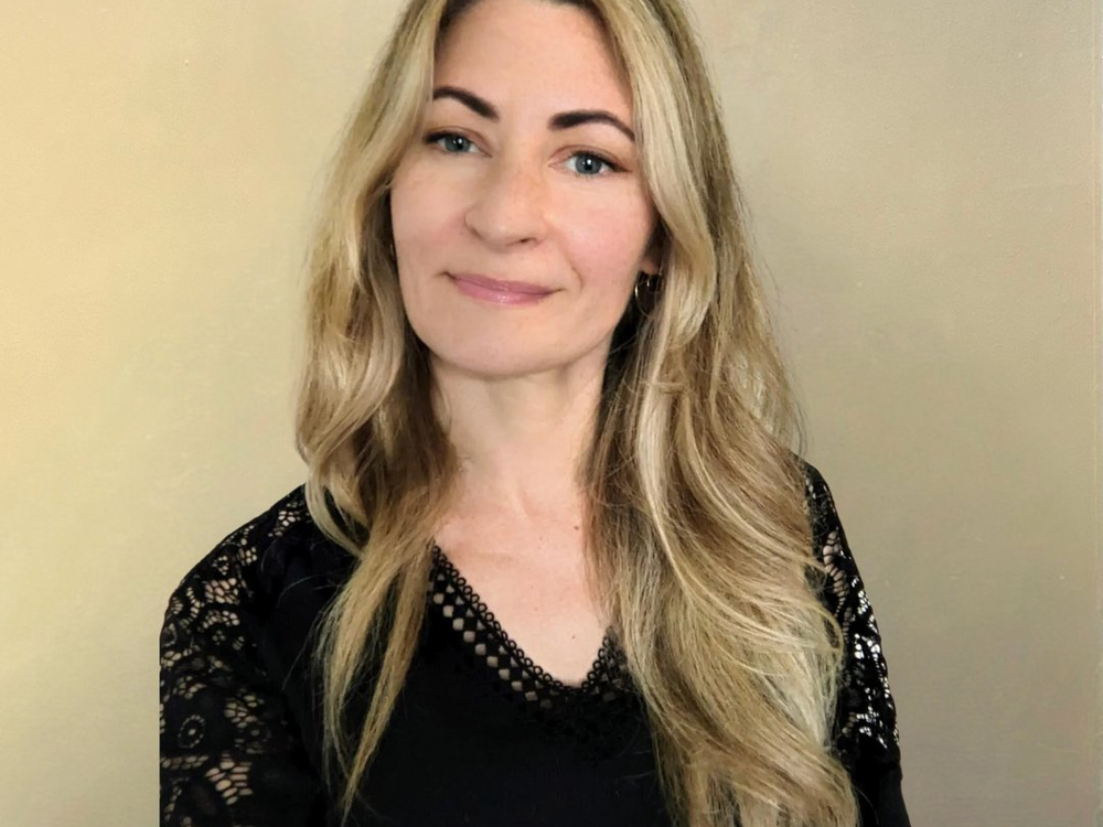 Woman with long blonde hair wearing a black lace top against a beige background
