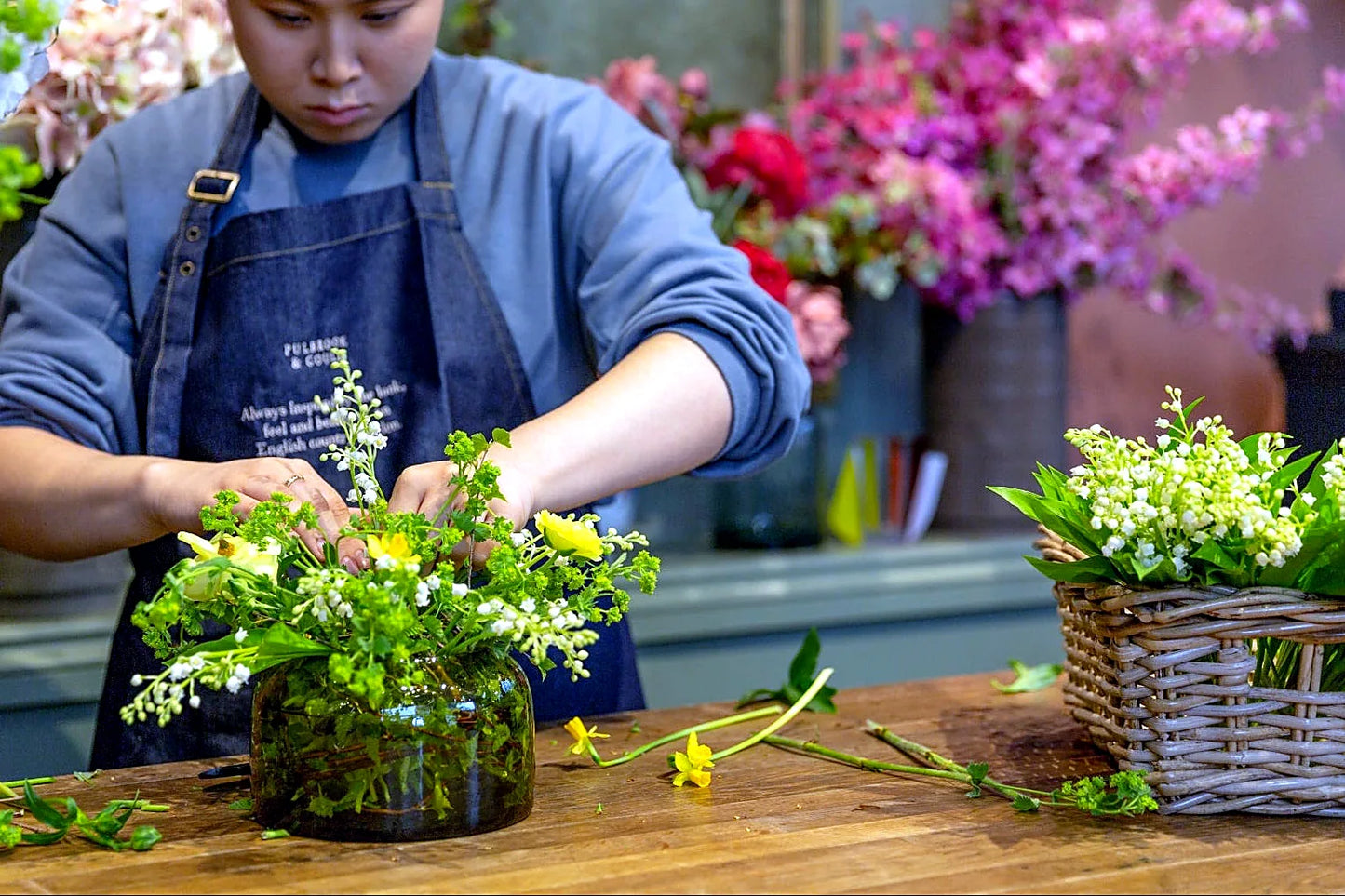 Gardener in denim apron arranging fresh green stems and small white flowers in a glass jar on a wooden table