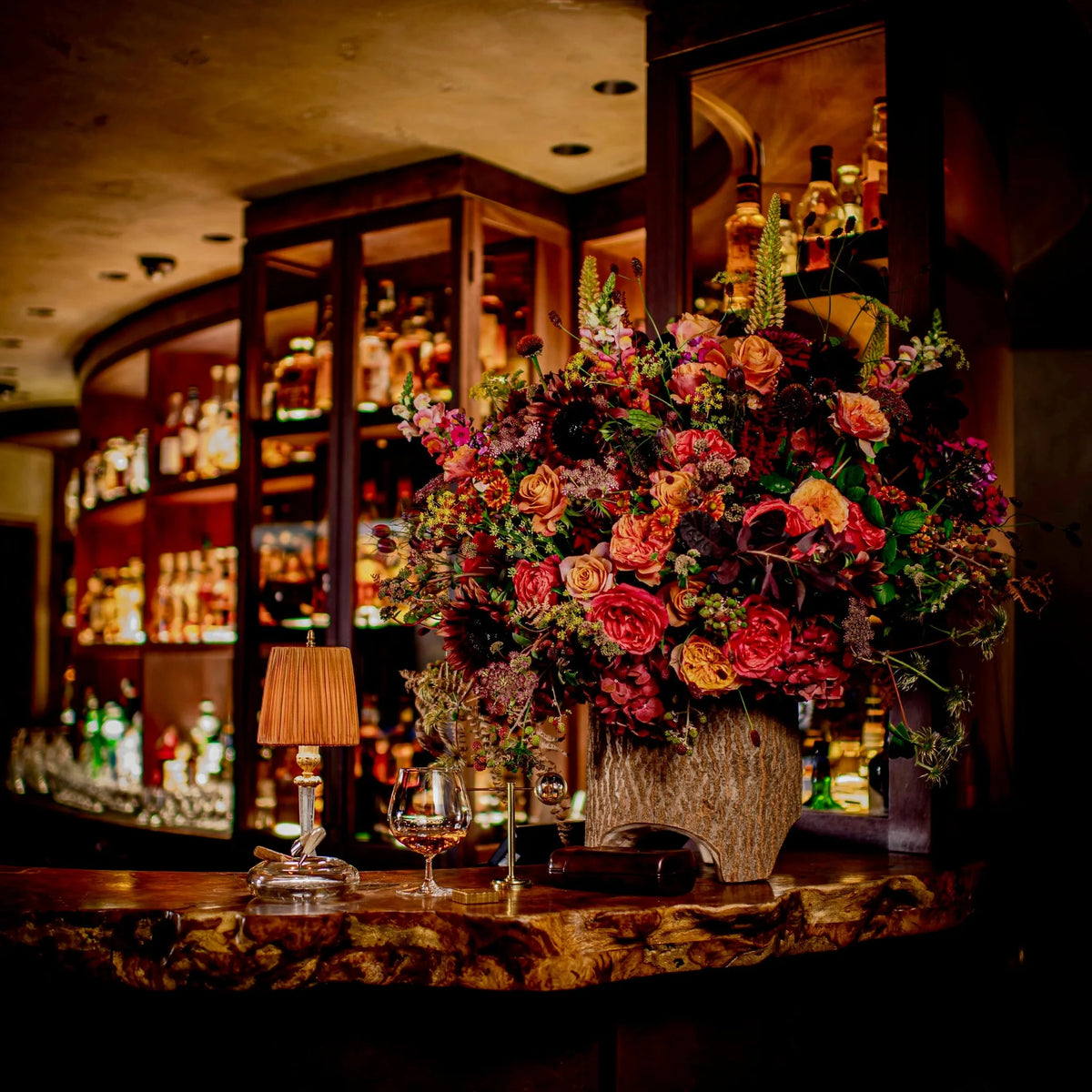 Vivid red and orange floral arrangement on a bar counter with backlit whisky bottles, corporate event, by Pulbrook & Gould