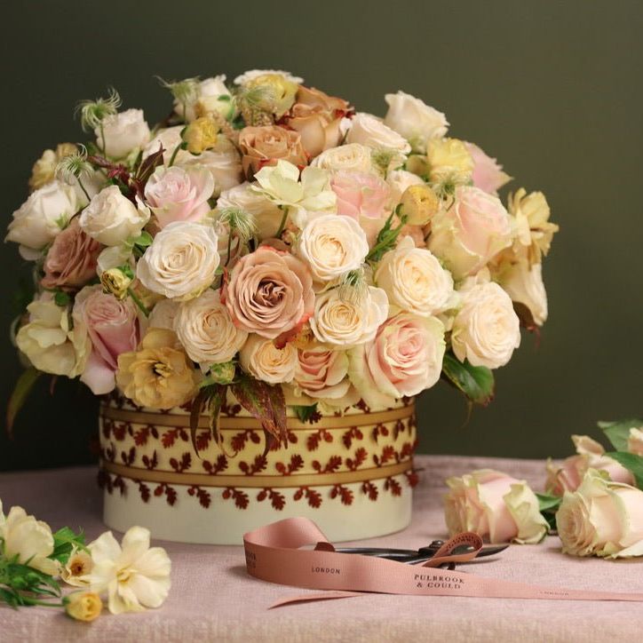 Elegant hatbox arrangement of blush, ivory and soft peach roses with ranunculus, styled in a decorative box with loose stems and ribbon displayed alongside.