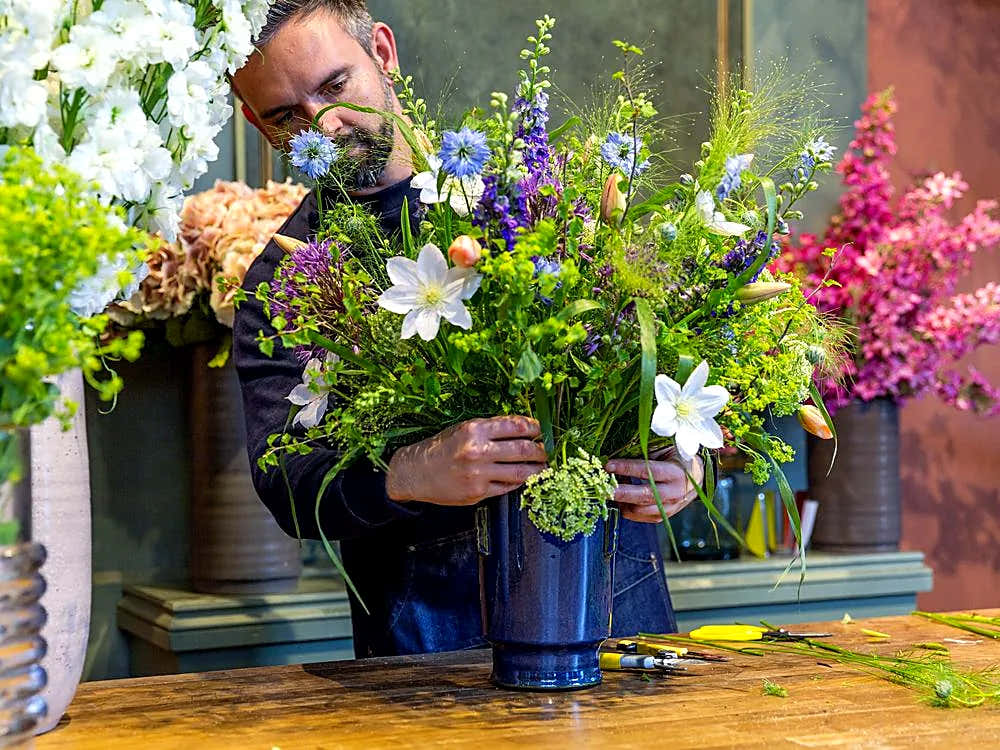 Florist arranging a colourful bouquet of wildflowers in blue vases, with a man holding stems at a workbench