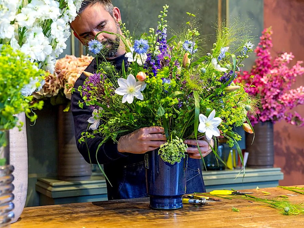 Florist arranging a colourful bouquet of wildflowers in blue vases, with a man holding stems at a workbench