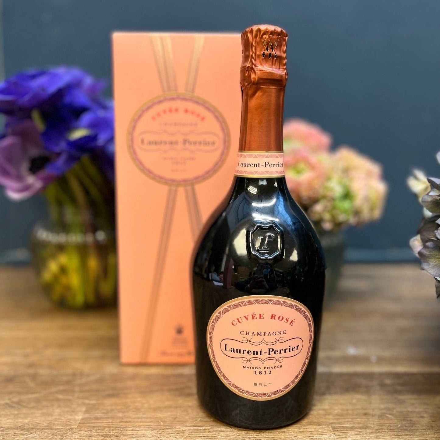 Bottle of Laurent - Perrier champagne with a pink label on a wooden surface, with flowers and a box in the background.