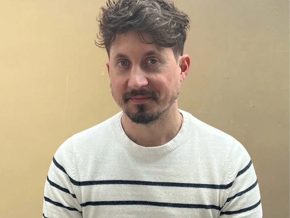 Man with short dark hair and a trimmed beard wearing a white striped jumper against a plain beige wall