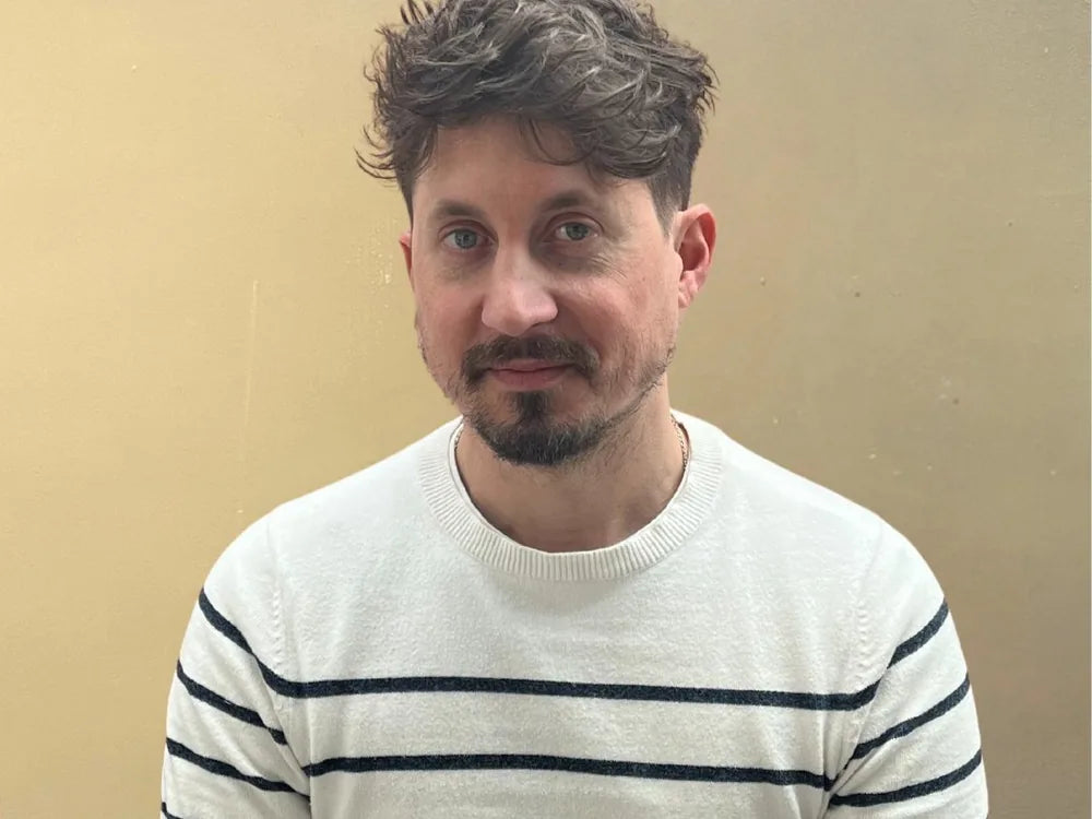 Man with short dark hair and a trimmed beard wearing a white striped jumper against a plain beige wall