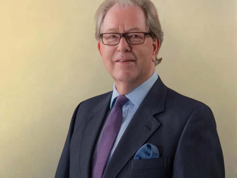 Man in a dark suit and purple tie with glasses, standing against a plain light background