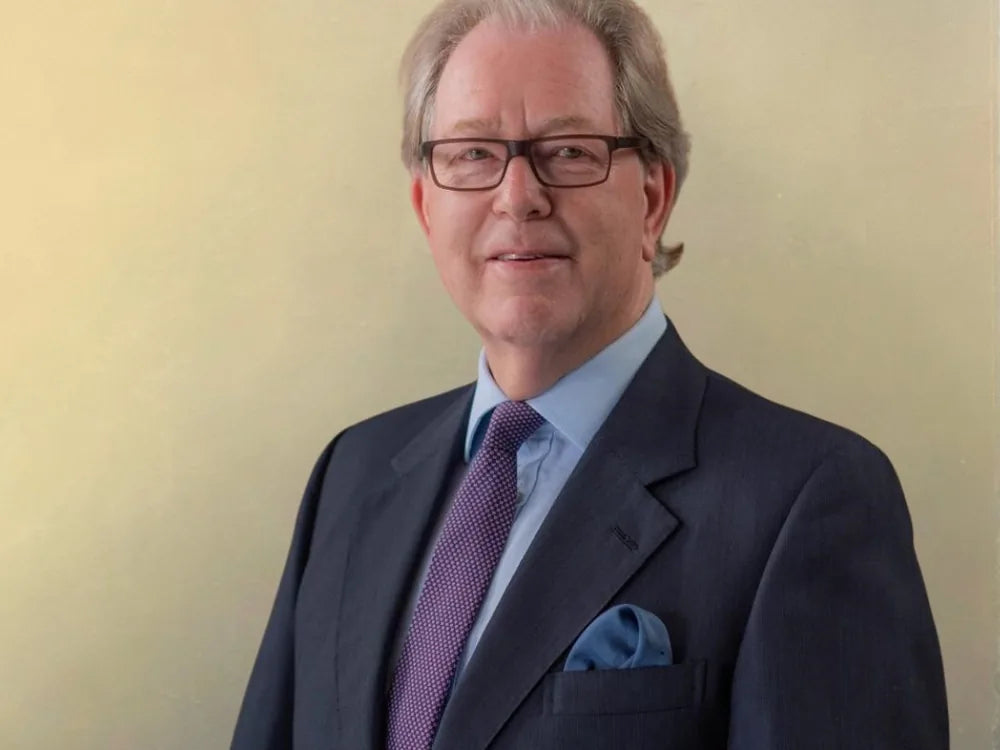 Man in a dark suit and purple tie with glasses, standing against a plain light background