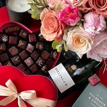 Overhead close-up of the Pulbrook & Gould Rosamund gift set showing a red heart-shaped box of luxury dark chocolates, a Bollinger Champagne bottle, a Pulbrook & Gould signature scented candle and blush and peach roses in a branded dark green box