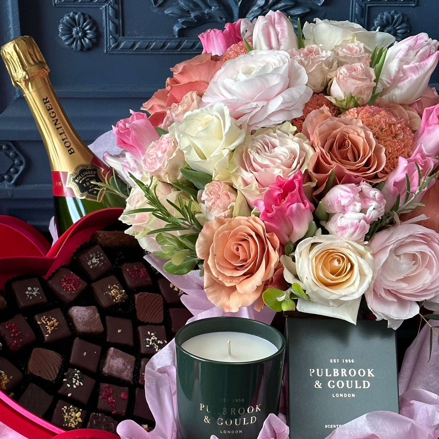 Pulbrook & Gould Rosamund gift set featuring a bouquet of blush garden roses, peach roses, pink tulips, white ranunculus and cerise carnations in a dark green branded box, alongside a Bollinger Champagne bottle, a red heart-shaped box of luxury chocolates and a Pulbrook & Gould signature scented candle against a dark panelled background