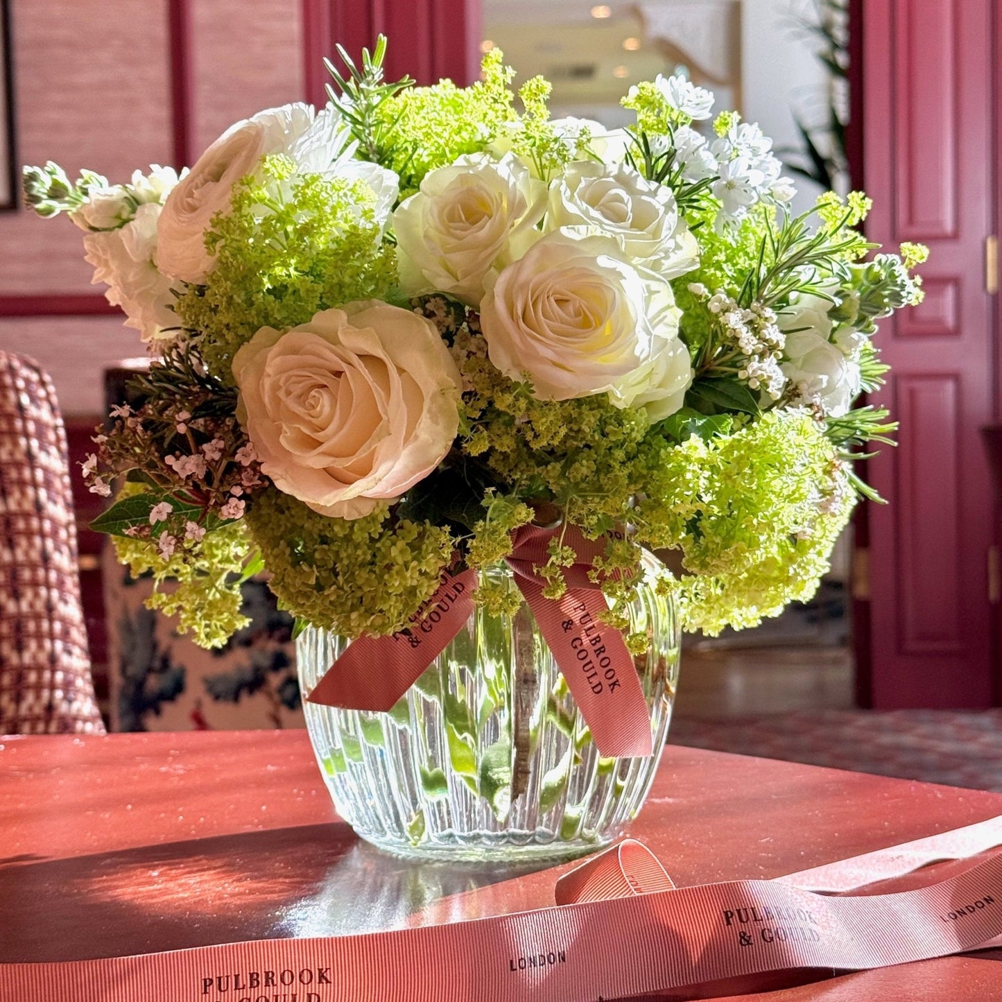 Bouquet of flowers in a clear vase with a pink ribbon on a red surface.