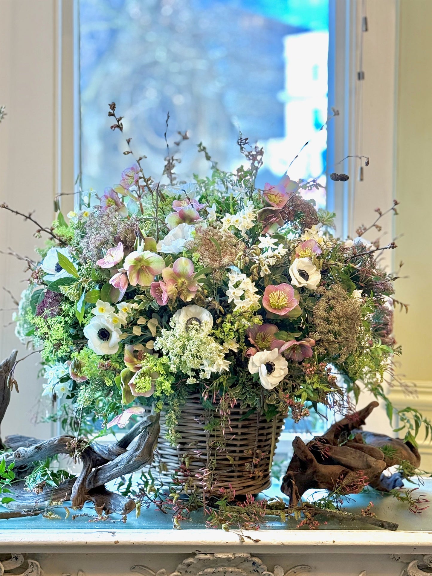 Floral arrangement in a woven basket on a decorative surface with a window in the background