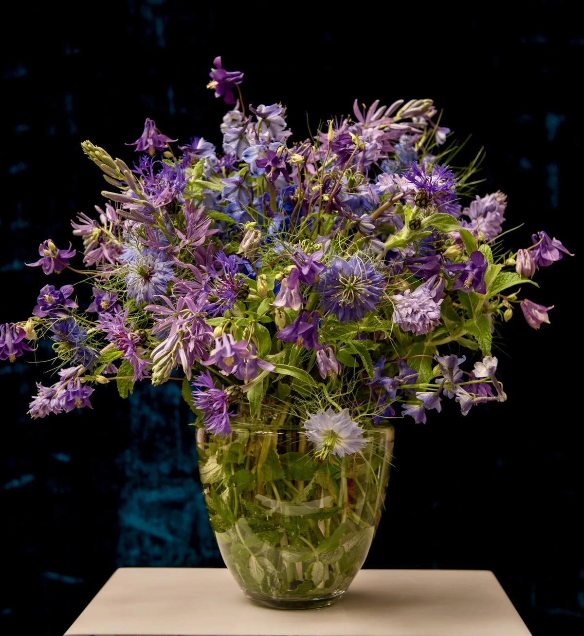 Wild-style purple and blue flower arrangement in a glass vase against a dark teal backdrop, weekly, by Pulbrook & Gould