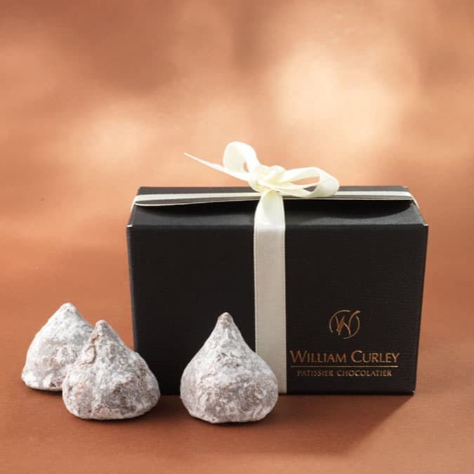 A black box of William Curley chocolates with a white ribbon, containing luxury Champagne Truffles, with two truffles in front of the box against a copper - colored background.