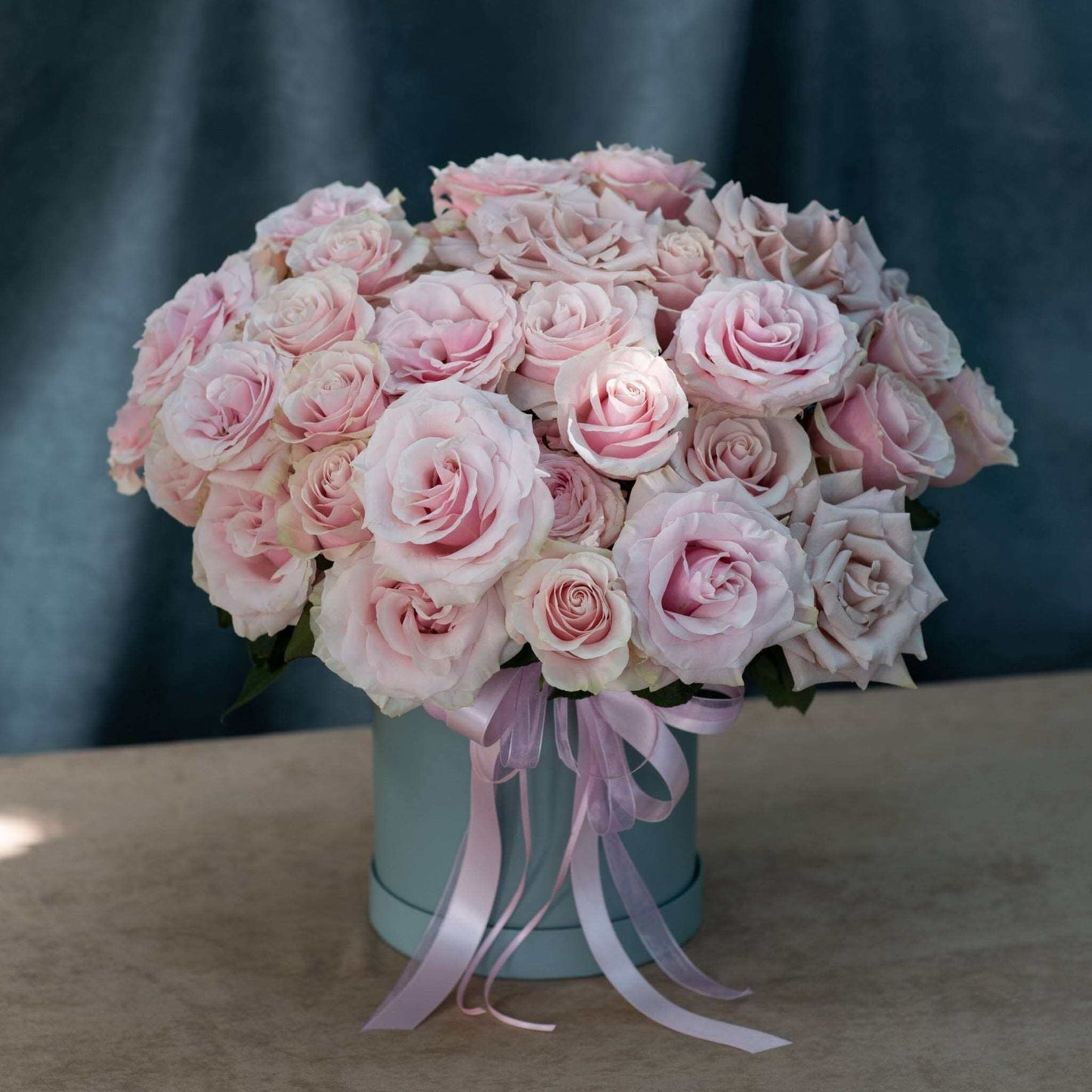 A hatbox arrangement of blush pink roses, tied with a pink ribbon, set against a blue background.